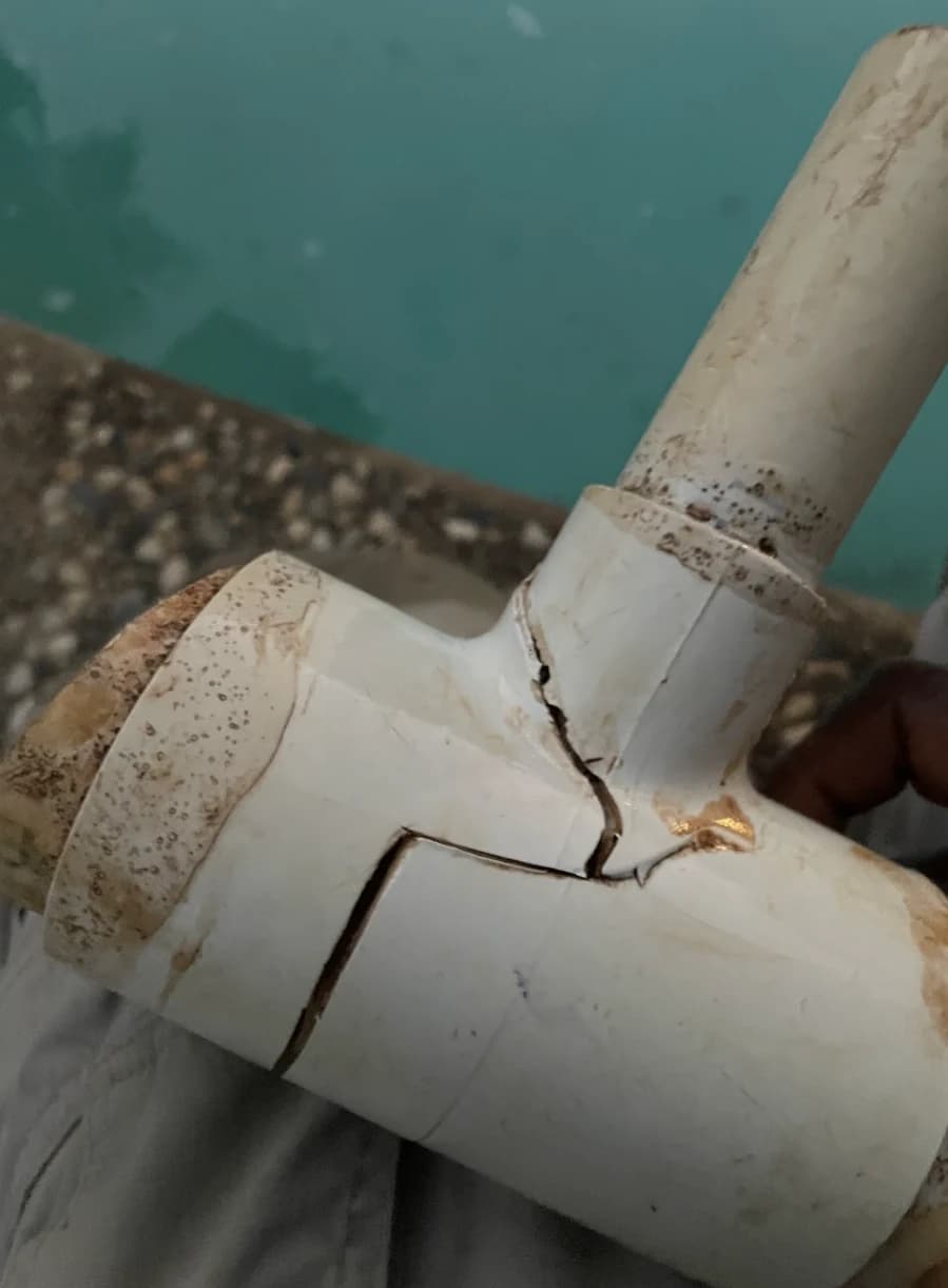 Cracked white PVC tee fitting held up beside a pool